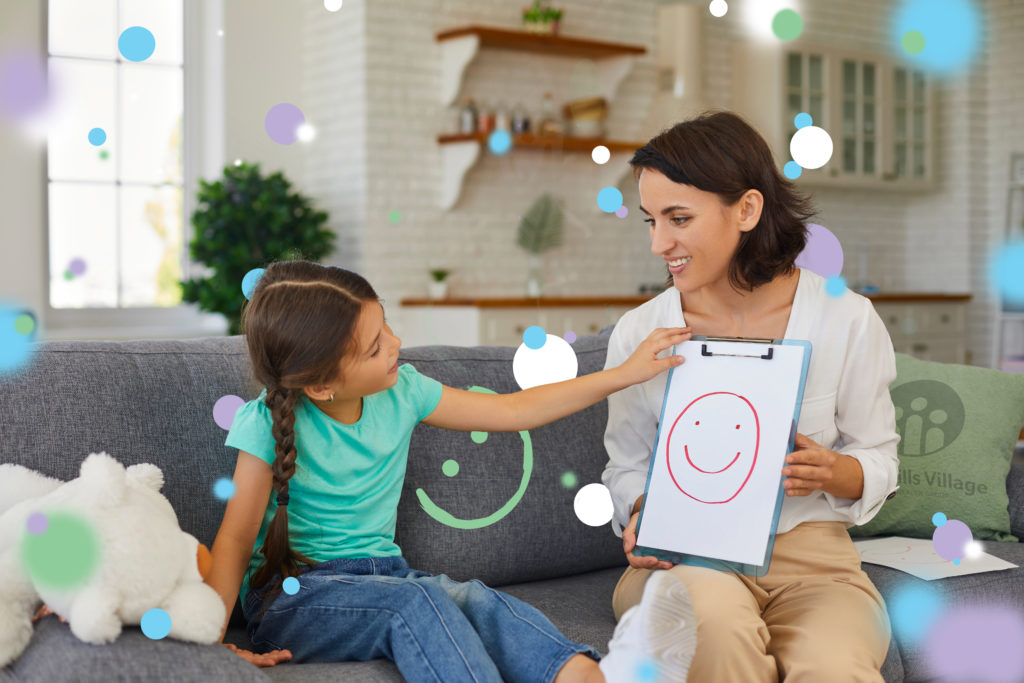Smiling assertive psychologist with picture of happy emoji talking about emotions with 8 - 10 year old girl. Supportive private therapist and child communicating during therapy session at home