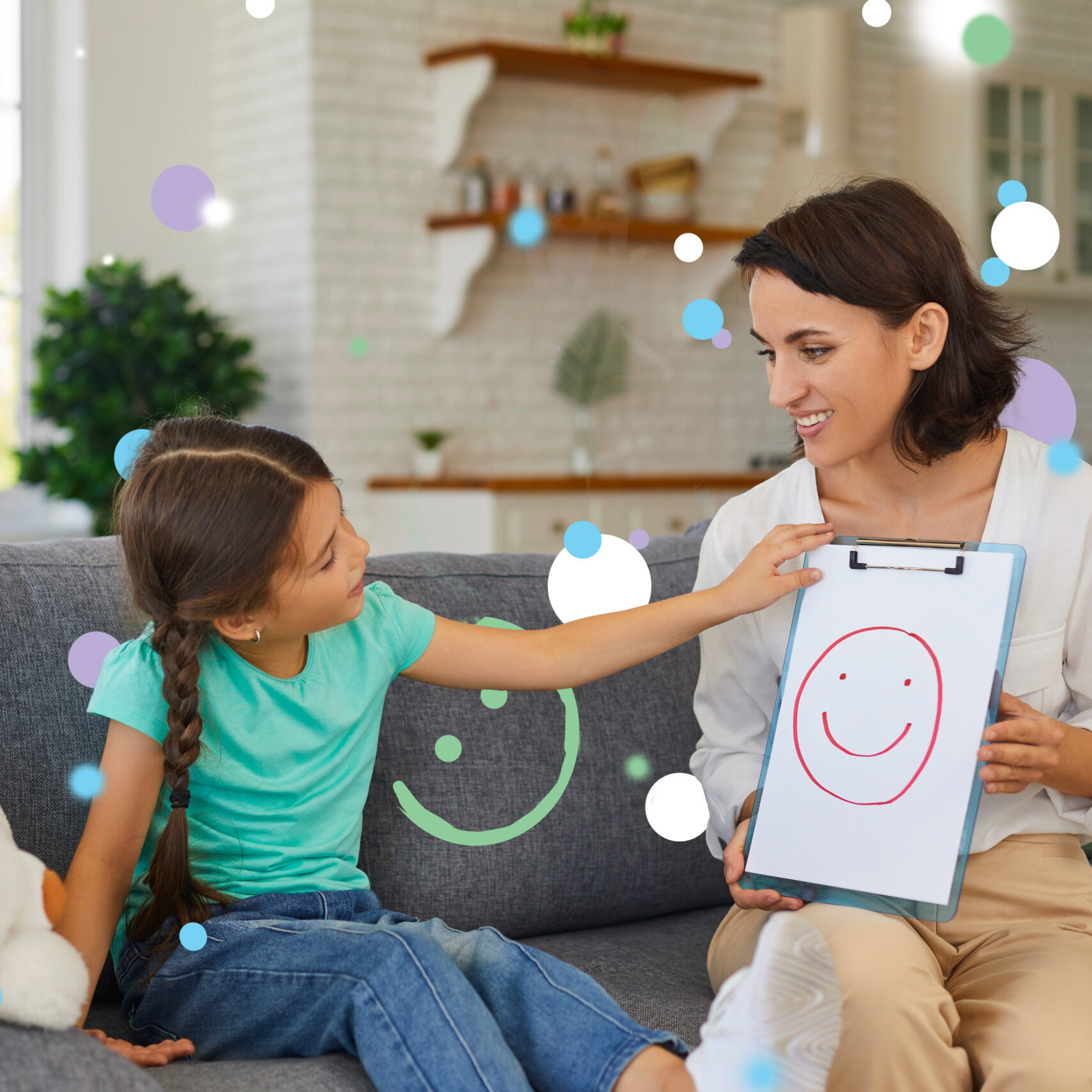 Smiling assertive psychologist with picture of happy emoji talking about emotions with 8 - 10 year old girl. Supportive private therapist and child communicating during therapy session at home
